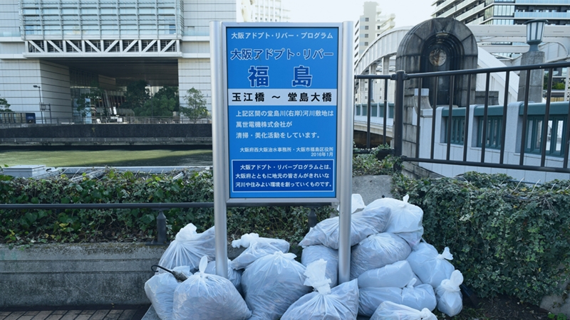 『河川清掃活動“アドプト・リバー・プログラム”』の看板前に集められたごみ袋